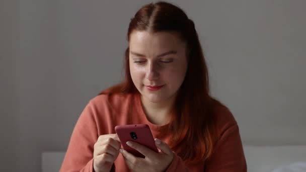 Téléphone. Jeune fille souriante du millénaire caucasien utilisant un smartphone. Jeune fille tenant un téléphone portable, bavarder dans les réseaux sociaux, assis sur le lit dans le salon. — Video