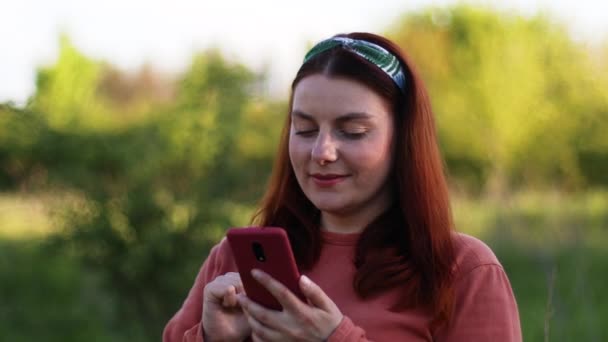 Femme positive utilise le téléphone mobile, lit des nouvelles dans le parc vert de la ville à l'été — Video