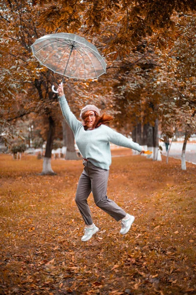 Vidám fehér lány ugrik fel egy átlátszó esernyővel az őszi parkban. Boldogság és szórakozás az esőben — Stock Fotó
