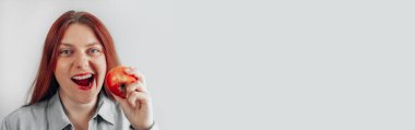 Gri arka planda kırmızı elma yiyen neşeli genç bir kadının portresi. Sağlıklı beslenme diyeti.