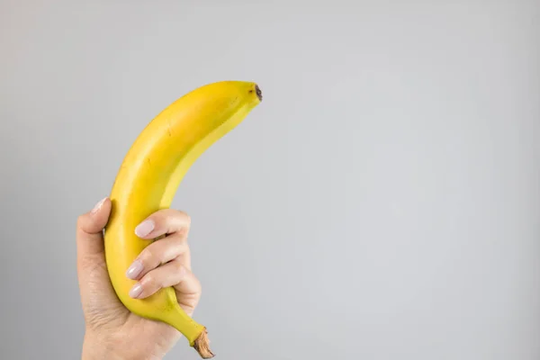 Mão feminina segurando uma banana amarela em fundo cinza colorido brilhante. Conceito de sexo seguro. — Fotografia de Stock