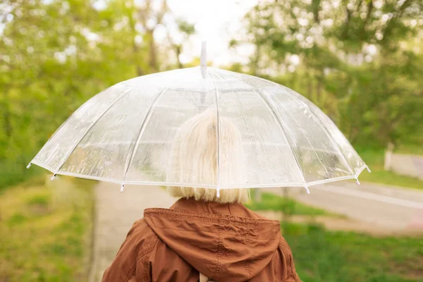 Mujer caucásica camina por calles húmedas y lluviosas bajo un paraguas transparente. Clima frío. Paraguas transparente húmedo en el clima lluvioso de otoño. Hermoso parque de otoño — Foto de Stock