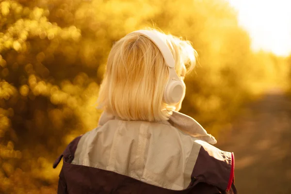 Güneşli bir günde şehir parkında yürürken kulaklıkla müzik dinleyen beyaz bir genç kadın. — Stok fotoğraf