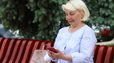 Orta yaşlı, gülümseyen sarışın kadın şehrin caddesinde yürürken akıllı telefon kullanıyor..
