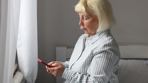 Adulto mujer seria está escribiendo un mensaje de texto, charlando, sentado en la cama. — Vídeos de Stock