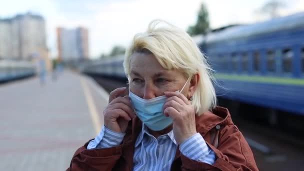 Volwassen blonde vrouw zet een beschermend medisch masker op haar gezicht op het perron van het station. Reizen per trein — Stockvideo