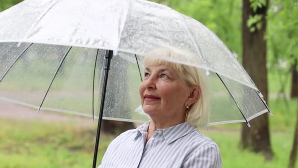 Volwassen senora 50 jaar oud vrouw met een transparante paraplu terwijl ze buiten staat te wandelen in het park en de tuin — Stockvideo