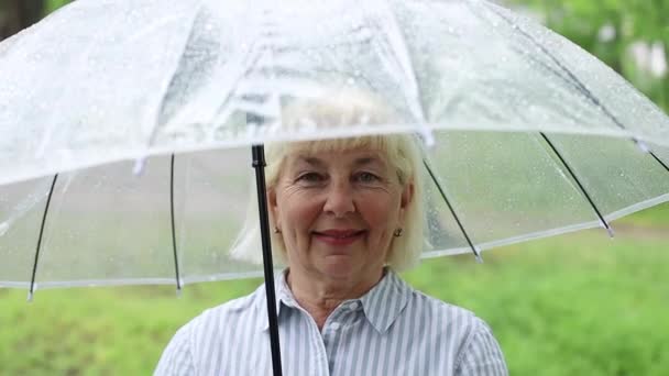 Volwassen senora 50 jaar oud vrouw met een transparante paraplu terwijl ze in de zomer buiten in het park en de tuin staat te wandelen — Stockvideo