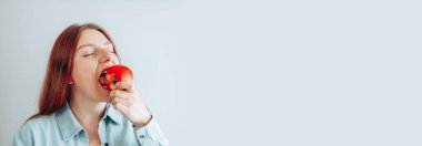 Gri arka planda kırmızı elma yiyen neşeli genç bir kadının portresi. Sağlıklı beslenme diyeti. Elma vitamini..