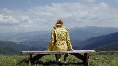 Yol kenarındaki bankta oturan parlak sarı ceketli genç bayan turistin arka görüntüsü güzel dağların manzarasının tadını çıkarıyor.