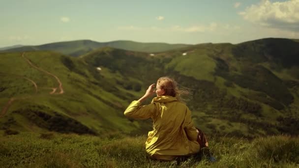 Gyönyörű szőke nő turista világos sárga dzsekiben ül a hegy tetején, és kinyújtott kézzel nézi Mountain Valley-t. Arany haj a friss levegőben szél — Stock videók