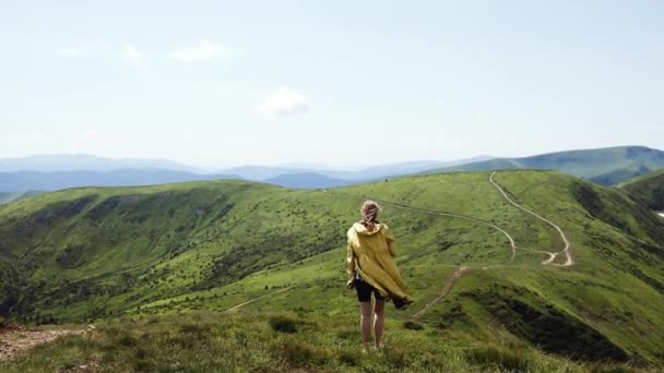 Mujer turista viajera en una chaqueta de color amarillo brillante caminando en la cima de la montaña en verano día soleado bajo la luz del sol. Hermosas montañas vista al paisaje. — Vídeo de stock