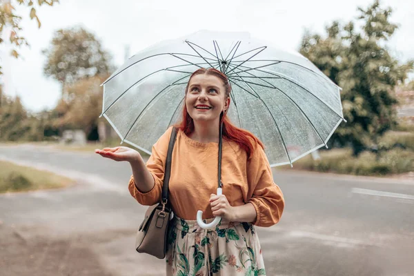 Uma linda garota fica sob um guarda-chuva transparente e estende a mão, pegando gotas de água. Banner horizontal, espaço de cópia — Fotografia de Stock