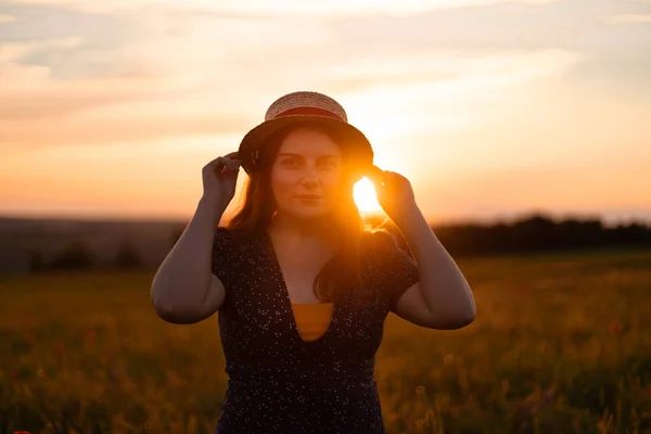 Belle fille aux cheveux roux avec un chapeau de paille et une robe d'été dans un champ aux rayons du coucher du soleil — Photo