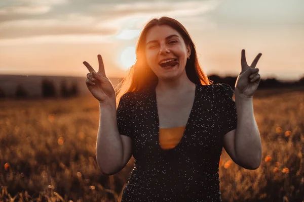 Ευτυχισμένο χαμογελαστό κορίτσι με καλοκαιρινό φόρεμα κοιτάζει την κάμερα και δείχνει το σύμβολο της ειρήνης με το δάχτυλό της στο ηλιοβασίλεμα — Φωτογραφία Αρχείου