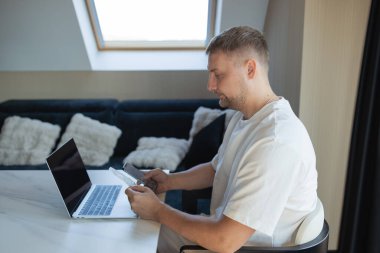 Genç adam bilgisayar ve telefonla masada oturuyor, faturaları ödüyor ya da internetten alışveriş yapıyor. Nakitsiz ekonomi, mobil bankacılık, ev bütçesi, mali teknoloji ve uzak çalışma.