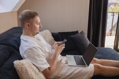 Kanepede dizüstü bilgisayarı ve akıllı telefonu olan rahat bir adam uzaktan çalışıyor, internetten alışveriş yapıyor ya da mali durumunu kontrol ediyor. Serbest çalışma kavramı, dijital yaşam tarzı, uzaktan çalışma, mobil bankacılık, online