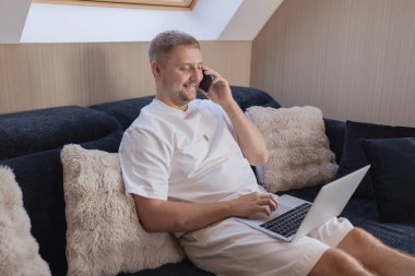 Kanepede dizüstü bilgisayarla oturan ve akıllı telefondan konuşan gülümseyen adam. Uzaktan çalışma, çevrimiçi iletişim, serbest çalışma, dijital yaşam tarzı, e-bankacılık, online alışveriş veya iş görüşmesi kavramı. Samimi