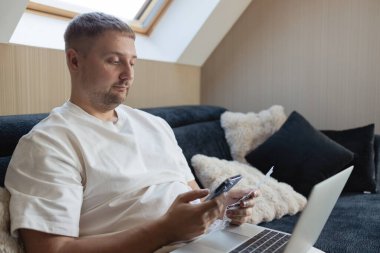 Yetişkin bir adam elinde dizüstü bilgisayar, akıllı telefon ve fatura ile kanepede oturuyor. Ev finansmanı ve internet ödemelerini yönetiyor. Dijital bankacılık, e-ticaret, fintech, bütçe, evden çalışma ve