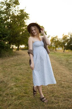 Altın saat boyunca kırsal bir yolda yürüyen açık renk elbiseli ve şapkalı bir anne bekliyor, gülümsüyor ve doğanın tadını çıkarıyor. Annelik yaşam tarzı, hamilelik sağlığı, sağlık ve aile kavramı