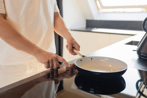 Крупный план человека в белой одежде, надевающего сковородку на электрическую плиту на современной кухне. Концепция приготовления пищи, повседневной жизни и домашнего образа жизни. Символ подготовки, комфорта, минималистичного интерьера