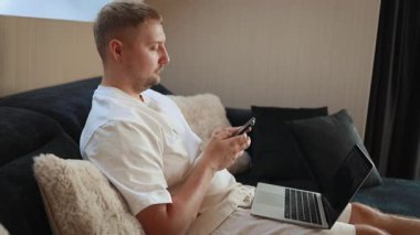 Evde oturan genç adam, güvenli online ödeme için dizüstü bilgisayar, akıllı telefon ve kredi kartı kullanıyor. Günlük yaşamda dijital bankacılık, uzaktan çalışma, finansal planlama ve modern teknoloji kavramı.