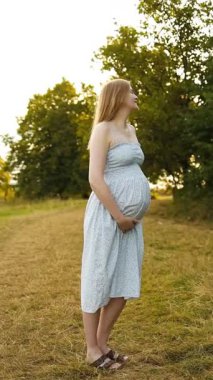 Akan yaz elbisesi içinde gülümseyen hamile bir kadın altın saatinde dışarıda duruyor, nazikçe karnındaki şişliği tutuyor. Annelik yaşam tarzı, hamilelik sağlığı, doğum öncesi bakım ve aile sevgisi kavramı