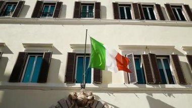İtalyan üç renkli bayrağı Roma 'daki tarihi bir hükümet binasının cephesinde zarafetle dalgalanıyor. Parlak güneş ışığı, ulusal gurur ve İtalyan mirasını sembolize ederek mimariyi aydınlatır..