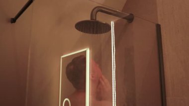 Sıcak ortam ışığıyla modern bir banyoda duş alan bir adamın videosu. Hijyen, rahatlama, kişisel bakım, ev hayatı ve günlük rutin kavramı. Sağlık, banyo tasarımı ve sağlık için ideal.