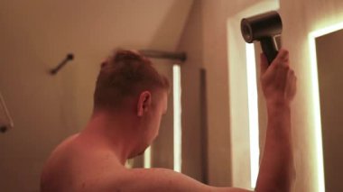 Sıcak ışıklandırmalı modern bir banyoda saç kurutma makinesi kullanan bir adamın videosu. Sabah rutini, kişisel bakım, hijyen ve evde kişisel bakım kavramı.