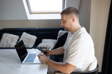 Evde oturan genç adam, güvenli online ödeme için dizüstü bilgisayar, akıllı telefon ve kredi kartı kullanıyor. Günlük yaşamda dijital bankacılık, uzaktan çalışma, finansal planlama ve modern teknoloji kavramı.