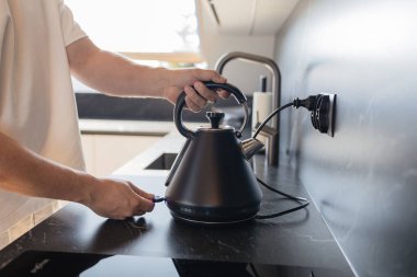 Modern mutfaktaki mermer duvarlı ocağa siyah elektrikli su ısıtıcısı koyan adamın yakın çekimi. Ev hayatı, yemek pişirme ve günlük rutin şeyler. Ev konforunun sembolü, şık iç mekan ve