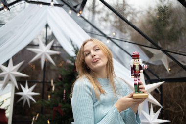 Açık mavi kazaklı gülümseyen sarışın kadın, Noel 'de içinde ışıklar ve süslemeler olan ahşap bir fındıkkıran oyuncağı tutuyor. Sıcak tatil atmosferi ve neşeli kış havası.