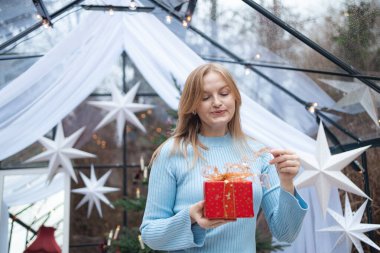 Açık mavi kazaklı, mutlu, genç, sarışın beyaz kadın altın kurdeleli kırmızı Noel hediye kutusunu açıyor. Noel ağacı, beyaz yıldızlar ve tatil ile samimi bir şenlik ortamı