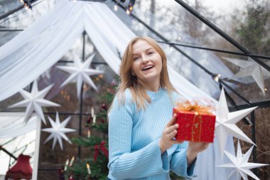 Açık mavi kazaklı, mutlu, genç, sarışın beyaz kadın altın kurdeleli kırmızı Noel hediye kutusunu açıyor. Noel ağacı, beyaz yıldızlar ve tatil ile samimi bir şenlik ortamı