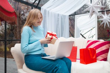 Açık mavi kazaklı sarışın kadın dizüstü bilgisayarlı rahat koltukta oturuyor, elinde kırmızı kurdeleli bir Noel hediyesi kutusu ile online video görüşmesi yaparken gülümsüyor. Kırmızı çantalı şenlikli arka plan