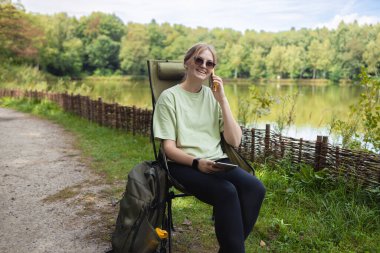 Göl kenarında sırt çantasıyla kamp koltuğunda oturan ve akıllı telefonuyla konuşan neşeli bir kadın. Dijital bağlantıyı, seyahat yaşam tarzını ve doğa eğlencesini temsil eden rahatlama sahnesi.