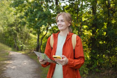 Sırt çantalı, turuncu ceketli, elinde kağıt harita ve akıllı telefon tutan genç bir kadın bir orman gölünün yakınında yürürken. Navigasyon, seyahat, macera, yürüyüş ve eko-turizm kavramı.