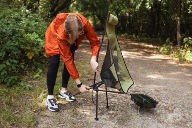 Turuncu ceketli birinin, orman yolunda katlanır bir kamp sandalyesini kurarken yakın çekimi. Yürüyüş hazırlığı, açık hava macerası, ekolojik seyahat ve doğada rahatlama kavramı.
