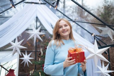 Açık mavi kazaklı, mutlu, genç, sarışın beyaz kadın altın kurdeleli kırmızı Noel hediye kutusunu açıyor. Noel ağacı, beyaz yıldızlar ve tatil ile samimi bir şenlik ortamı