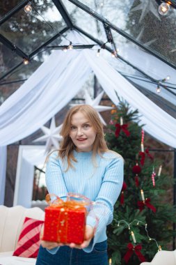 Açık mavi kazaklı neşeli sarışın kadın kırmızı bir Noel hediyesi kutusu tutuyor ve gülümsüyor. Noel ışıkları, ağaç ve süslemelerle süslü kış arka planı rahat bir ortam yaratıyor.