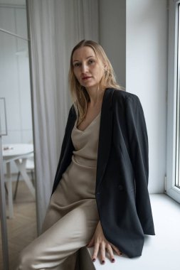 Bej bir elbise giyen zarif bir kadın ve siyah ceket parlak minimalist bir iç mekanda pencerenin kenarında oturuyor. Doğal gün ışığı modern moda, güven ve zamansız stili vurgular. Sakin ol nötr tonlar ve