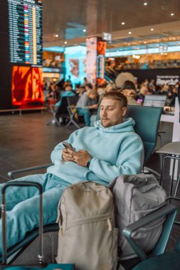 Neşeli genç beyaz adam havaalanında oturur ve telefonundaki bir uygulama ile bir sonraki uçuşu kontrol eder. Uçağı bekler ve seyahat ederken çalışır. Yüksek kalite fotoğraf