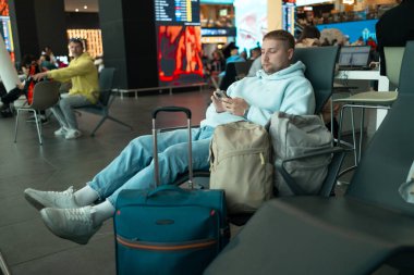 Havaalanı kapısında oturan genç adam uçuş için bekliyor, akıllı telefon ve bavulla uçaklara bakıyor. Modern hava yolculuğu konsepti. Yüksek kalite fotoğraf