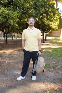 Yeşil bir parkta elinde sırt çantası olan yetişkin bir adamın tam vücut portresi. Günlük yaşam tarzı, seyahat ve eğlence konsepti. Erkek turist ya da şehir gezgini şehir parkında eğleniyor.