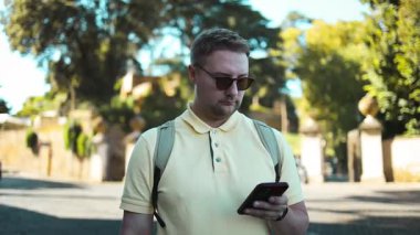 Güneşli bir günde açık havada yürürken akıllı telefon kullanarak güneş gözlüğü ve sırt çantası takan sıradan bir yetişkin. Ağaçları ve tarihi mimarisi olan bir Avrupa şehrinde kentsel yaşam sahnesi