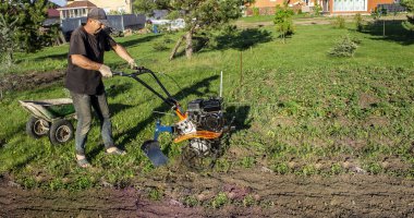 Baharda bir erkek, iş kıyafetleriyle toprağı sürüyor. Motoblock marka bir traktörü var.