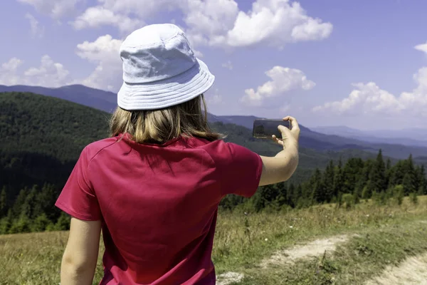 Kırmızı tişörtlü ve mavi şapkalı bir kız akıllı telefonuyla dağ manzarasının fotoğrafını çekiyor.
