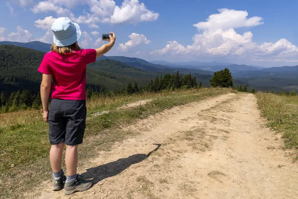 Kırmızı tişörtlü ve mavi şapkalı bir kız akıllı telefonuyla dağ manzarasının fotoğrafını çekiyor.