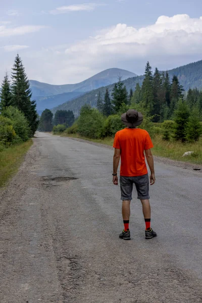 Yeşil şapkalı ve turuncu tişörtlü güçlü bir adam yolda dikilip dağa bakıyor. Hoverla Dağı 'na giden yolun başlangıcı, Karpatlar, Ukrayna